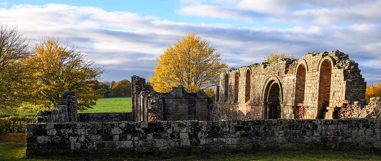 The roofless sandstone chapel of White Ladies Priory with round-headed Norman arches, behind a low stone wall and framed by golden autumn trees