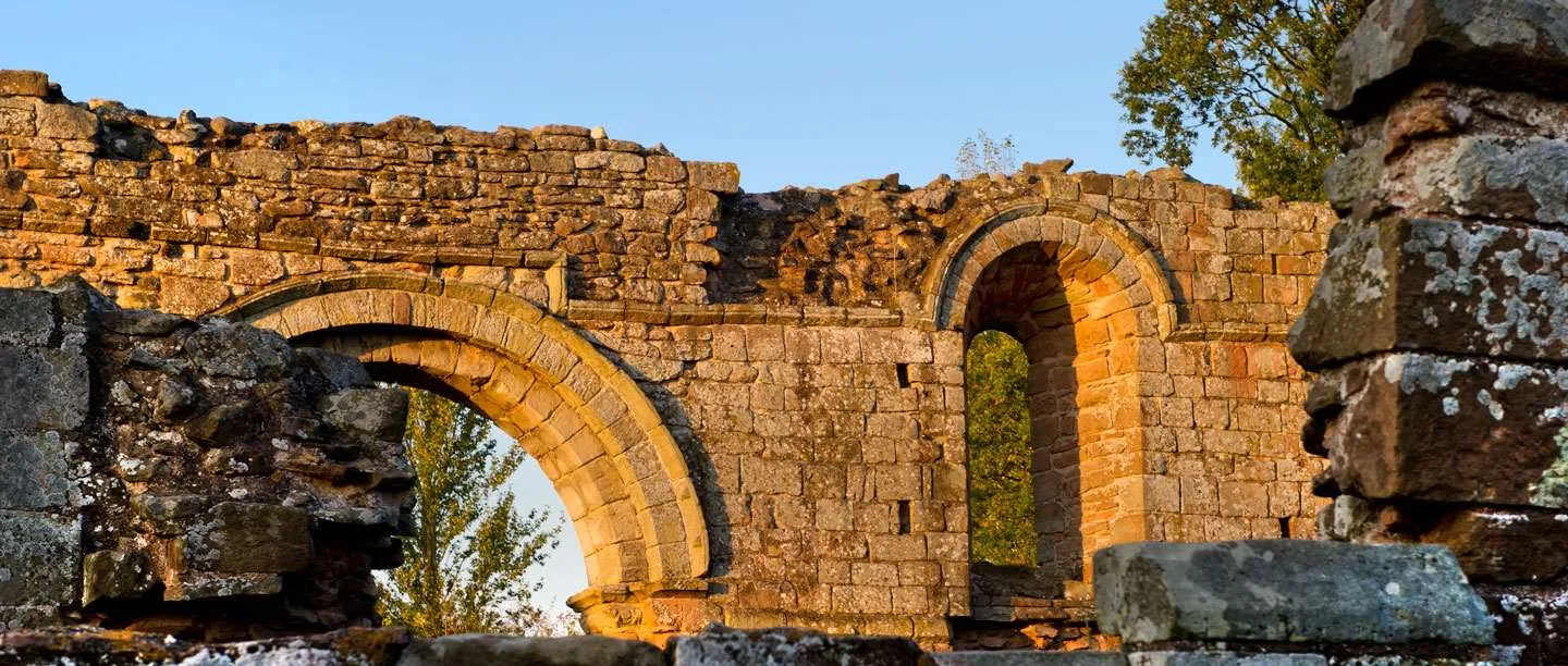 Round Norman arches in the warm sandstone walls of White Ladies Priory, lit by low evening sun against a blue sky