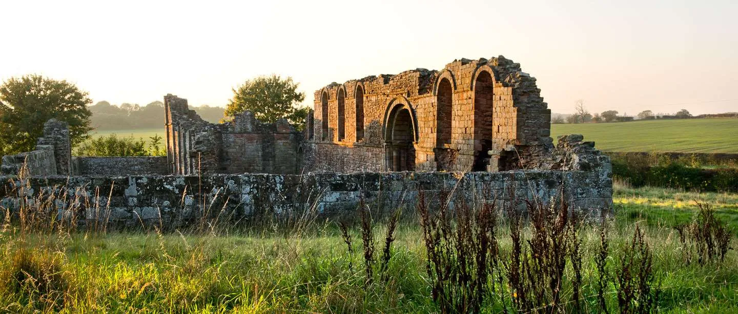 The ruined chapel of White Ladies Priory with tall round-arched windows glowing in early morning light, behind a low wall and long grass