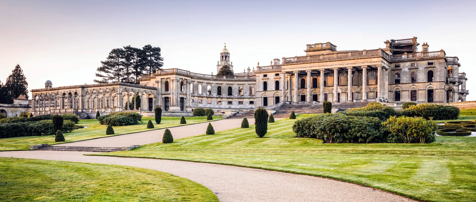The ruined mansion's portico and the long arcaded conservatory above lawns and clipped yews at dusk